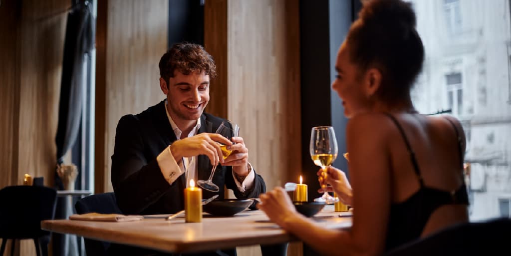 A well-dressed couple on a date smile and swirl white wine glasses at a candle-lit dinner table after meeting each other through a matchmaker.