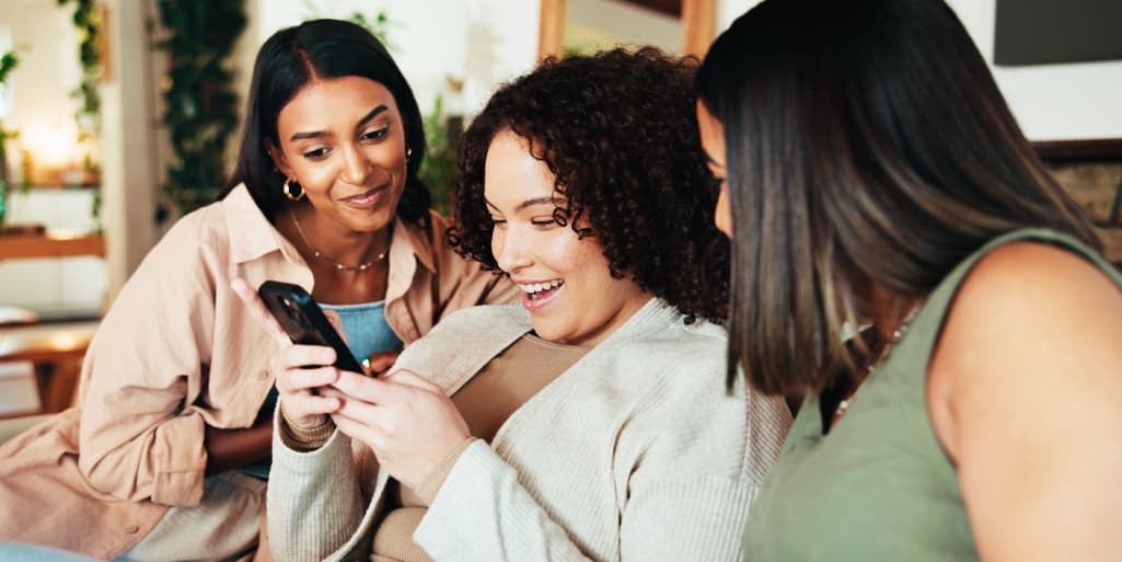 A woman looks excitedly at her phone while her two female friends help her choose a date on a dating app.